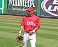 Baseball player standing in the outfield holding his glove with his right hand and wearing a red jersey that says Phillies, white wristband on his left hand and a red baseball cap.