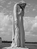 A statue of a mourning cloaked young woman. In one hand she has a bunch of laurel and holds her chin in the other hand.