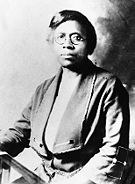 An African American female with short dark hair, round eye glasses, and a solemn expression sitting down and facing the camera.
