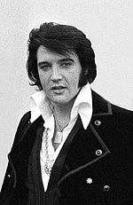 Posed photo of Elvis, shoulders and chest, wearing a shirt with a large, white collar and a coat with big lapels, medallions around his neck.