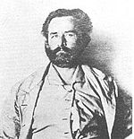 Grainy black and white photograph showing the face and torso of an about 50-year-old bearded man looking into the camera. He is wearing a jacket, and collared shirt that appears to be buttoned only at the top.