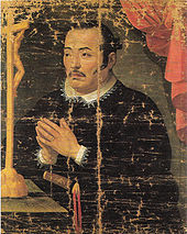 Painting of an Asian man in black priest robes with white collar praying in front of a crucifix.