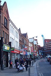 A row of shops facing onto a very busy pavement. A large six-storey brick building is visible in the background
