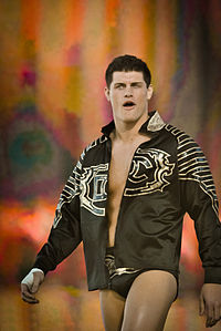 A caucasian male with black hair is wearing short black wrestling tights and a black jacket featuring a gold design on both sleeves and on the front. He has white tape on one of his hands.