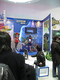 Inside a large, brightly lit convention center room with white walls is positioned a promotional display booth for a video game. A saleswoman clad in a blue shirt and skirt and a red bowtie motions towards several illustrations on the booth, explaining their implications. The illustrations are anime-styled and depict several outlandish and brightly colored creatures. Three men in dark jackets, one at a laptop, watch the demonstration.