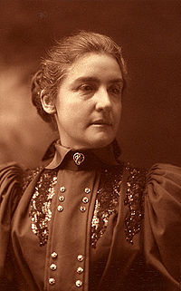A black and white photograph featuring a woman wearing an old-fashioned dress and with hair drawn back into a bun.