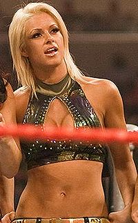 A blonde Caucasian woman wearing a green crop top standing in a wrestling ring. A red ring rope is visible in front of the woman.