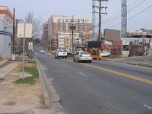 Ground-level view of a mid-sized street in an urban area, with one lane closed in the direction headed away from the viewer.  In the far background are high-rise office and residential buildings, while the near background features a construction site with a large crane against an overcast sky on a winter day.