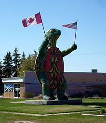 A large turtle statue standing on two legs and holding a Canadian flag in one hand an American flag in the other.