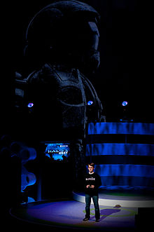 A man stands on a large stage, talking to the audience. Behind him is a flatscreen television displaying the game's title menu; behind that is a large representation of an ODST.