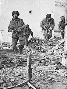 Four men in a bombed and gunshot building, walking towards the camera over rubble