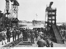 A large ship slides into the water. A tall crane stands on one side and a large scaffolding on the other. A crowd of spectators has gathered all around.