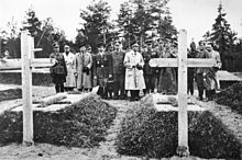 17 men, most in military uniform, stand in a cemetery, viewing two graves.