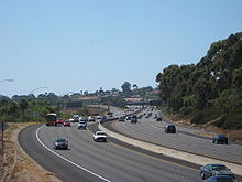 Under clear skies, vehicles travel on the freeway. Brush surrounds the freeway, but buildings can be seen in the distance.