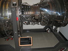 An engine is shown on a display stand at a museum, with the front of the engine facing left. Sections of the casing are cut away and replaced with clear plastic, revealing the booster blades, the compressor blades, and the turbine blades, from left to right.