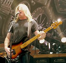 A fifty-eight year old man is singing into a microphone while playing a bass guitar. Behind him are stage lights and a partial view of a large steam train. His long white hair partly obscures his face. He wears a dark tee-shirt and darker pants.