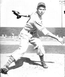 A man wearing a baseball uniform stands on the pitcher's mound having just thrown a baseball.