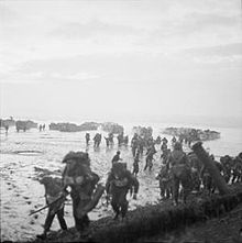 Men in the foreground burdened with equipment, behind them are mud or sand flats and in the distance landing craft
