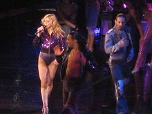 A female blond performer singing in a purple leotard and high heeled shoes. She is singing while looking to the right while on her left, a group of dancers in violet colored clothes strike different poses.