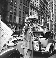 A man in a big-city street between parked cars holds a folded newspaper up in front of his face with one hand, and carries other copies with his other hand. The man's suit and the cars' styles are from the 1930s. The newspaper masthead is "Social Just..." and the huge lead headline reads "ANNIVERSARY OF VERSAILLES ... THREAT TO U.S. PEACE".