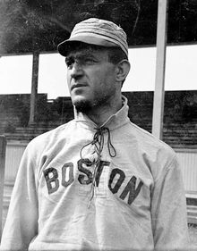 A man wearing a baseball cap and jersey with "Boston" written across the chest shown from the waist up looks to the left.