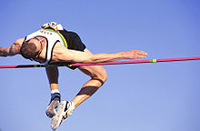 A man in an athletic uniform is jumping over the high jump bar headfirst and backwards. His legs trail behind his body as he clears the bar.