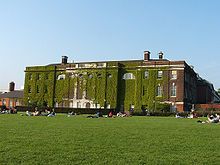 Massive, rectangular, three-storey brick building, covered in ivy. People are sitting in groups on the large front lawn.