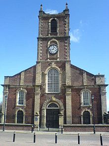 A brick church with stone dressings seen from the south.  The west tower has a clock and pinnacles, and along the south face of the body of the church are Georgian-style windows