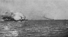A ball of flame engulfs a large gray warship. Several smaller ships are in the distance.