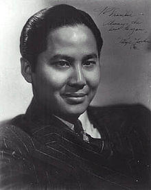 Head and shoulders publicity shot of a young, smiling man, short hair slicked back and wearing a suit and tie.