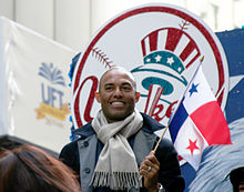 Mariano Rivera wearing a dark pea coat and gray scarf smiles while holding a red, white, and blue flag. He stands in front of a red, white, and blue logo that reads "Yankees".