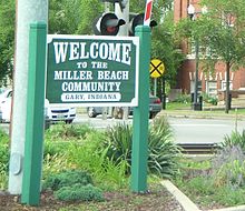 A large sign reading "Welcome to the Miller Beach Community, Gary, Indiana," next to a railroad crossing on a street with cars passing.