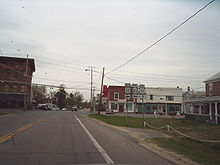 A two-lane highway approaches an intersection with another highway regulated by a blinking signal. Small, mostly two-story buildings surround the rural junction. A sign assembly indicates that NY 22 northbound and US 11 southbound turn left while US 11 northbound is straight ahead.
