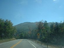 A two-lane highway in a forested area. To the right of the highway is a sign indicating that a hill is ahead. In the distance is a large mountain completely covered with trees.