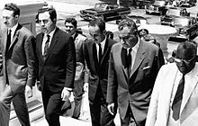 Five men wearing suits and ties walking up the steps of a building. Behind them are three other men, a large stone fountain, and a parking lot with four cars visible