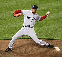 man wearing a grey baseball uniform that says "BOSTON" in navy letters clutches a baseball behind his head with his left hand as he prepares to throw it.
