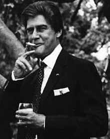 A middle-aged man of European descent smiles while holding a glass in his left hand and retrieving a cigarette from his mouth with his right. He is immaculately turned-out in a dark-coloured suit and looking to the viewer's left.