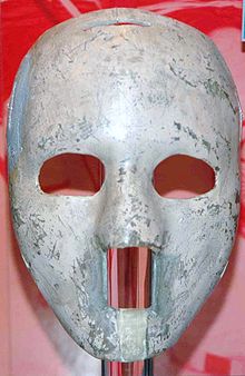 the mask is white and of solid construction, with egg-sized oval cutouts for the eyes and a rectangular cutout from the base of the nose to below the lower lip