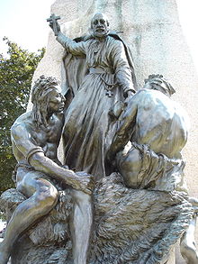 A bronze monument featuring a Christian missionary holding a cross high over the heads of two North American aboriginal peoples sitting at his feet