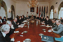 Thatcher is the only woman in a room, where a dozen men in suits sit around an oval table. Reagan and Thatcher sit opposite each other in the middle of the long axis of the table. The room is decorated in white, with drapes, a gold chandelier and a portrait of Lincoln.