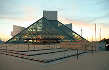 An extended glass pyramid in front of a tall grey building with strangly shaped outcroppings