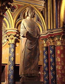 Full statue of a slender middle-aged man, standing on a pedestal in a chapel. The man is smooth-skinned, clean-shaven, has wavy neck-length hair, and is standing in a faded blue robe. The forearms of the statue are missing.