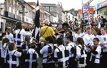 A street lined with shops is filled with hundreds of people. In the foreground are children wearing black vests each one defaced with a large white cross. The children surround a fiddler. In the background are spectators.