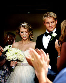 A blond teen female wearing white wedding gown is smiling while looking down. She is standing beside a blond man in a black tuxedo, who is also looking down. Both their arms are intertwined. A group of people is seen clapping while looking at the couple.