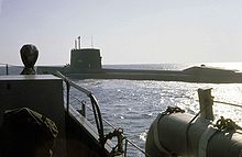 Color photograph depicting the port side of a large surfaced nuclear submarine taken from the bow of an escorting surface ship as the submarine leaves port.