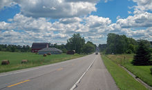 A road under a blue sky with white clouds goes past a farm on the left with rolls of hay in its fields and a red barn. In the distance the road climbs a small rise