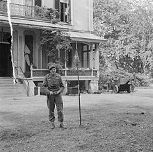 Man standing in front of imposing house with a pennant of a flag pole beside him