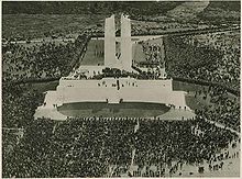 A memorial ceremony. Thousands of people are surround the monument on all sides. A crowd of people are also standing on the main platform of the memorial.