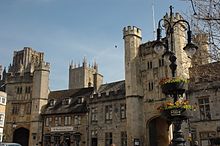 Ornate stone buildings. 2 archways beneath towers. In the foreground is a lamp stand with flowers.