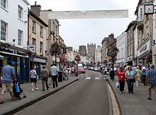 Street scene with shops on either side of the road and many pedestrians on the pavements. In the distance is the tower of the cathedral.